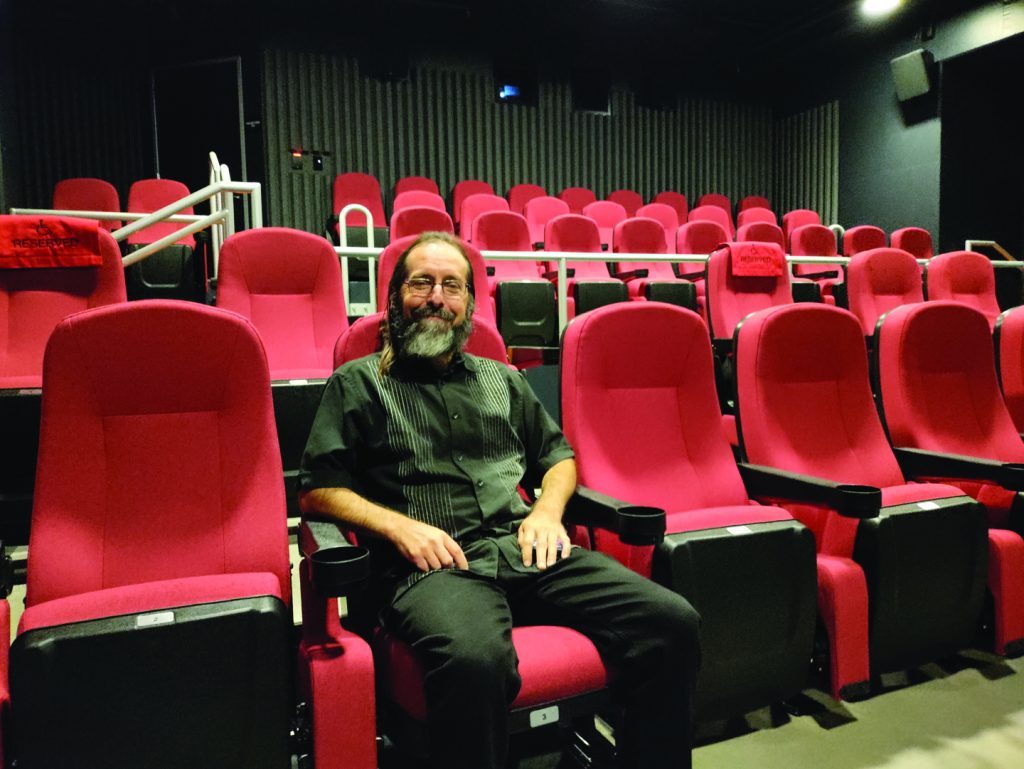 A man with a long gray and black beard wearing all black sits in a theater full of red seats.